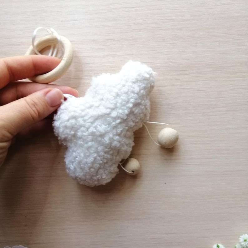 Pode incluir: Brinquedo de pel&uacute;cia branco e felpudo em forma de nuvem com contas de madeira penduradas. O brinquedo est&aacute; preso a um anel de madeira.