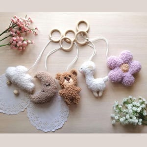 Puede incluir: Un móvil para bebé con cinco juguetes de peluche: una nube blanca, una luna creciente marrón, un oso marrón, una llama blanca y una flor morada. Los juguetes están unidos a anillos de madera y cuelgan de cuerdas blancas.