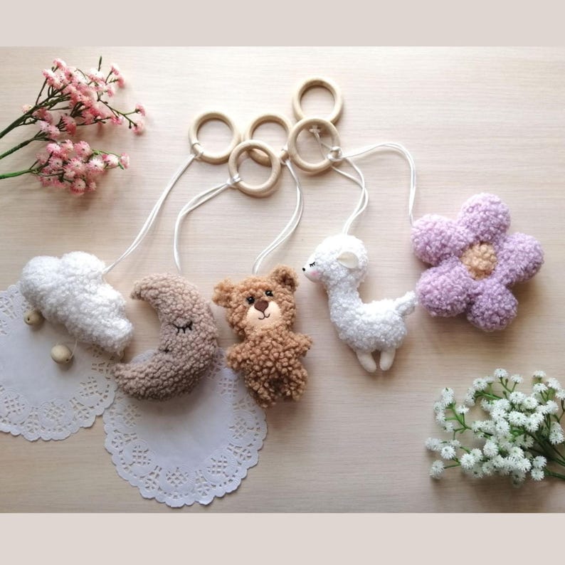 Pode incluir: Um m&oacute;bile para beb&ecirc; com cinco brinquedos de pel&uacute;cia: uma nuvem branca, uma lua crescente marrom, um urso marrom, uma lhama branca e uma flor roxa. Os brinquedos s&atilde;o presos a an&eacute;is de madeira e pendurados em cordas brancas.