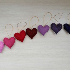 May include: A set of eight felt heart ornaments in a gradient of pink, red, and purple hues. Each heart has a gold string attached for hanging.