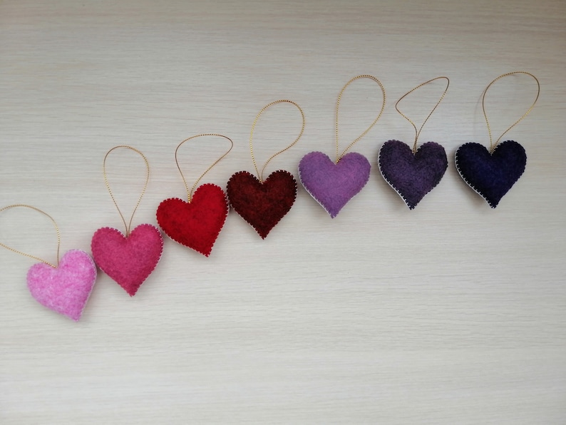 May include: A set of eight felt heart ornaments in a gradient of pink, red, and purple hues. Each heart has a gold string attached for hanging.