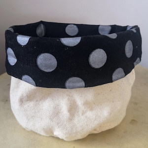 Peut inclure: Un panier de rangement en toile blanche avec une doublure en tissu à pois noirs et argentés. Le panier est rond et a une large ouverture.