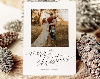 May include: A Christmas card with a family photo, featuring a family of four in a field. The card has the words "merry christmas" in elegant script, with "from the Johnsons" below. Decorative silver trees and pinecones are in the background.