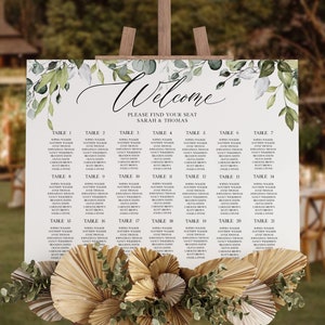 Peut inclure: Un plan de table de mariage avec un motif floral vert et blanc. Le plan est intitulé "Bienvenue" et liste les tables avec les noms des invités. Le plan est affiché sur un chevalet en bois avec un arrangement décoratif en forme d'éventail de feuilles de palmier séchées et de verdure.