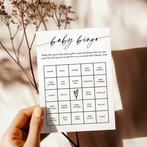 May include: A white baby bingo card with black text. The card has a grid of 25 squares with different baby-related items listed in each square. The items include diaper, bowl, onesie, baby shoes, baby carrier, baby blanket, car seat, diaper bag, bouncer, baby carrier, lotion, rattle, bath towel, stroller, baby playmat, diaper cream, baby powder, high chair, pillow, baby clothes, bath tub, baby wipes, burp cloth, and socks.