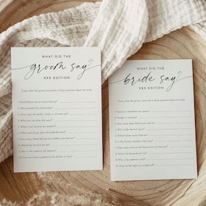 May include: Two white cards with black text on a wooden surface. The cards read "WHAT DID THE groom say?" and "WHAT DID THE bride say?" with a list of questions below. The cards are for a wedding game.