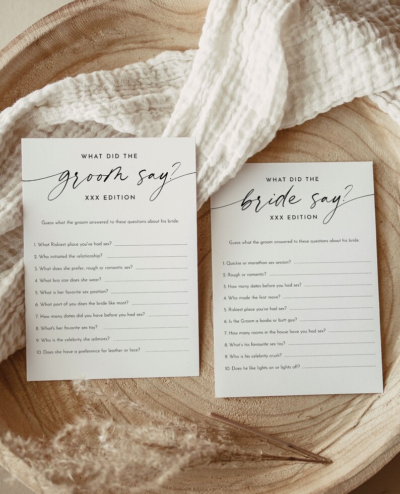 May include: Two white cards with black text on a wooden surface. The cards read "WHAT DID THE groom say?" and "WHAT DID THE bride say?" with a list of questions below. The cards are for a wedding game.
