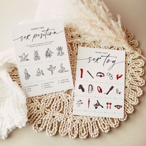 May include: Two white cards with black text and illustrations for a bachelorette party game. One card says "GUESS THE sex position" and the other says "NAME THAT sex toy". The cards are on a beige woven placemat.