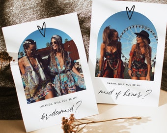 May include: Two white cards with a photo of two women in each. The cards have a blue and white arch with a heart at the top. The text on the cards reads "Amanda, will you be my bridesmaid?" and "Sarah, will you be my maid of honor?"