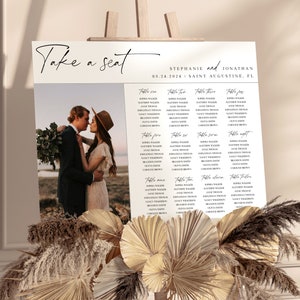 May include: A wedding seating chart with a rustic theme. The chart is printed on white paper and features a handwritten font. The chart is decorated with a photo of the bride and groom, as well as a floral arrangement. The chart lists the names of the guests and their assigned tables. The text on the chart reads "Take a seat", "Stephanie and Jonathan", "09.24.2024", "Saint Augustine, FL", and "Table one", "Table two", "Table three", "Table four", "Table five", "Table six", "Table seven", "Table eight", "Table nine", "Table ten", "Table eleven", "Table twelve".