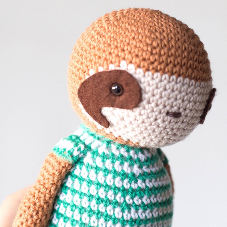 Puede incluir: Un juguete de peluche de perezoso tejido a crochet con pelaje marr&oacute;n, cara blanca y ropa a rayas verde y blanca.