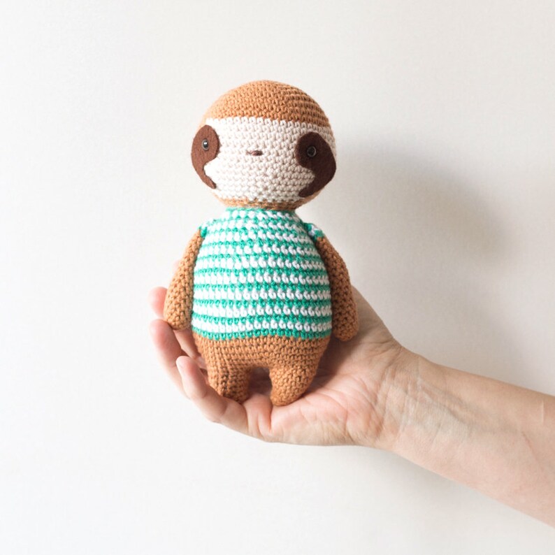Puede incluir: Un juguete de peluche de perezoso tejido a crochet con una camisa a rayas verdes y blancas. El perezoso tiene ojos marrones y una nariz marr&oacute;n.