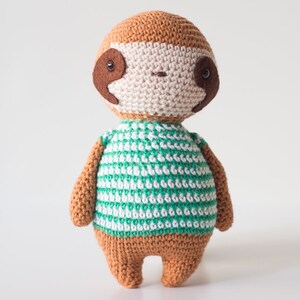 Puede incluir: Un juguete de peluche de perezoso tejido a crochet con una camisa a rayas verdes y blancas. El perezoso tiene pelo marr&oacute;n, una cara beige y ojos negros.