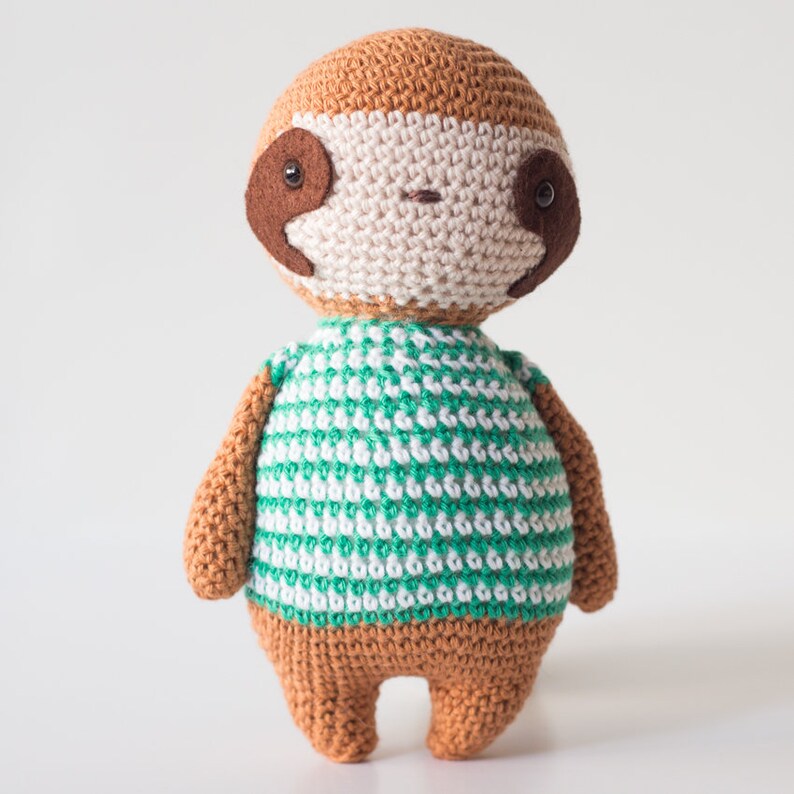 Puede incluir: Un juguete de peluche de perezoso tejido a crochet con una camisa a rayas verdes y blancas. El perezoso tiene pelo marr&oacute;n, una cara beige y ojos negros.