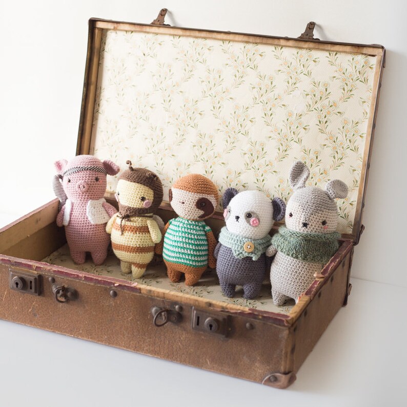 Puede incluir: Cinco animales de peluche tejidos a crochet, incluyendo un cerdo, una abeja, un perezoso, un panda y un conejito, est&aacute;n dispuestos dentro de una maleta vintage abierta con un forro de estampado floral.