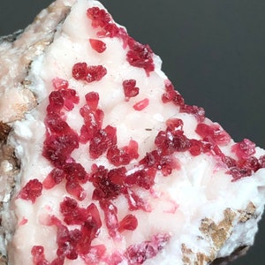 Puede incluir: Primer plano de un espécimen mineral con una base rosa claro y grupos de formaciones cristalinas de color rojo intenso. Los cristales están agrupados en la superficie, creando una textura. El fondo es gris oscuro.