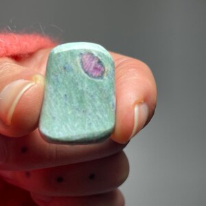 May include: A rectangular, polished gemstone with a light green base color and a vibrant pink inclusion. The stone is held in a hand wearing a coral-colored sweater. The gemstone's surface has a speckled texture.