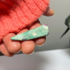 May include: A small, green, triangular-shaped stone with white and red inclusions. The stone is held in a hand, with a coral-coloured jumper in the background. The stone has a polished, smooth surface.