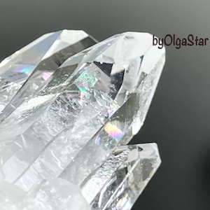 Pode incluir: Close-up de um aglomerado de cristais de quartzo transparente. Os cristais têm pontas afiadas e exibem arco-íris internos. A imagem é bem iluminada, destacando a clareza e as facetas do quartzo. O texto "byOlgaStar" é visível no canto superior direito.