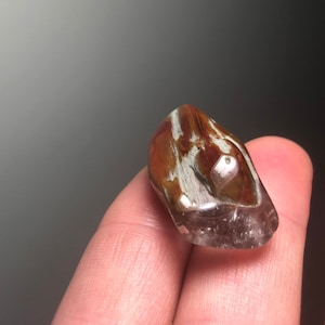 Pode incluir: Uma pedra preciosa polida, castanha e branca, com uma aparência translúcida. A pedra tem uma forma lisa e arredondada e é segurada em uma mão. A pedra preciosa tem uma mistura de cores castanhas e brancas.