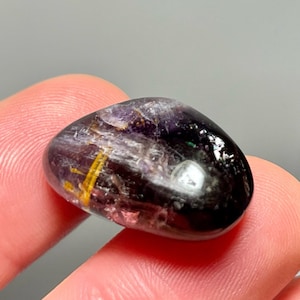 Pode incluir: Uma pedra preciosa polida, de forma oval, com uma mistura de cores. A pedra tem uma superfície escura e brilhante com raios roxos, dourados e pretos. O texto "byOlgaStar" é visível no fundo. A pedra preciosa é segurada entre dois dedos.