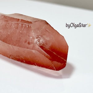 May include: A reddish-pink crystal with a faceted shape and a translucent quality. The crystal has a natural, rough texture and is set against a white backdrop. The image includes the text "byOlgaStar".
