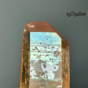 Può includere: Un grande cristallo sfaccettato con una calda tonalità marrone e riflessi iridescenti blu e verdi. La superficie del cristallo ha un aspetto strutturato e la parte superiore è tagliata con una faccia piatta e angolata. Il testo "byOlgaStar" è visibile.