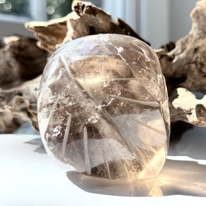 May include: A clear, polished quartz crystal carving shaped like a rounded leaf with etched veins on its surface. The stone is translucent, showing internal fractures and light reflections, and is positioned in front of a piece of driftwood.