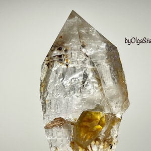 Pode incluir: Um cristal de quartzo transparente com uma ponta, apresentando inclusões internas e um depósito mineral amarelo dourado. O cristal está sobre um fundo branco, com o texto "byOlgaStar" visível.