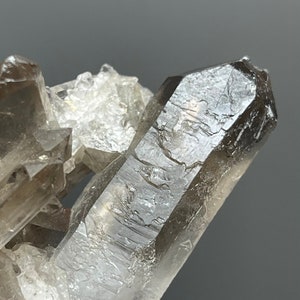 Pode incluir: Um aglomerado de cristais de quartzo fumê. Os cristais são de uma cor marrom esfumaçado translúcida com pontas afiadas e bem definidas. O quartzo é colocado sobre uma base cristalina mais clara, criando um contraste de cor e textura.