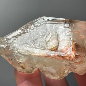 Puede incluir: Un cristal translúcido con bordes ásperos y un tono ámbar claro. El cristal tiene una apariencia en capas con fracturas internas y una textura ligeramente rugosa. La forma general es irregular, con algunos bordes afilados y una apariencia mayormente opaca.