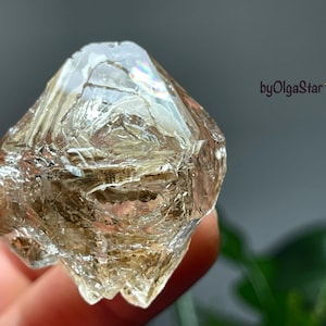 Puede incluir: Cristal facetado y translúcido con un tono marrón claro. La superficie del cristal tiene un aspecto texturizado, con patrones e inclusiones internas visibles. La imagen está sobre un fondo borroso, con el texto "byOlgaStar" visible.