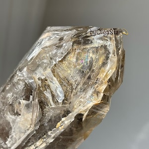 May include: Close-up of a large, translucent crystal with a rough, layered texture. The crystal has a light brown color with areas of iridescence, reflecting a rainbow of colors. The text "byOlgaStar" is visible on the top right.