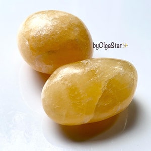 May include: Two smooth, oval-shaped yellow calcite stones. The stones have a polished appearance and a warm, golden hue. The text "byOlgaStar" is visible in the upper right corner.
