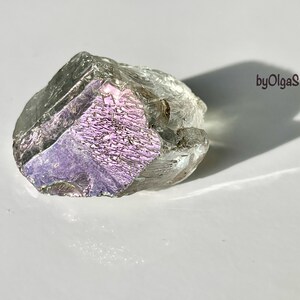 May include: A raw, iridescent crystal with a clear, gray exterior and a vibrant, purple and pink interior. The crystal is rough-hewn and has a textured surface. The image includes the text "byOlgaStar".