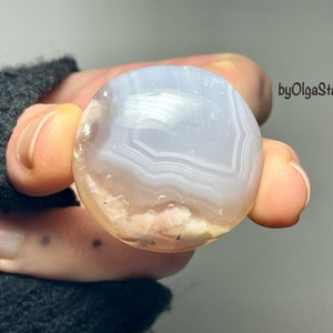 Puede incluir: Una gema redonda y pulida con un patrón en capas y bandas en tonos blancos, azul claro y melocotón. La piedra se sostiene en una mano, mostrando su superficie lisa y sus colores naturales. El texto "byOlgaStar" es visible.
