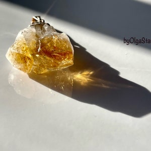 Op de afbeelding: Een ruwe, goudgele citrien kristallen hanger met een zilveren beugel. De ruw geslepen edelsteen wordt verlicht door zonlicht en werpt een schaduw. De afbeelding bevat de tekst "byOlgaStar".