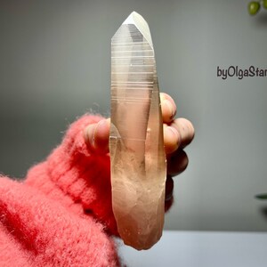 Puede incluir: Un cristal claro y alargado con una punta. El cristal tiene un tono marrón claro y parece estar sostenido en una mano. El fondo es gris suave, con un toque de follaje verde visible en el lado derecho. El texto "byOlgaStar" es visible.