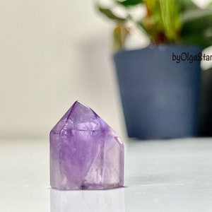Puede incluir: Una punta de cristal de amatista facetada. El cristal púrpura está cortado en una forma geométrica con una base plana y una parte superior puntiaguda. El cristal es translúcido, permitiendo el paso de la luz. El fondo está borroso, con una planta en una maceta azul.