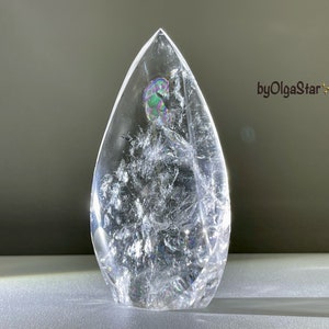 May include: A clear, teardrop-shaped crystal with internal fractures and iridescent inclusions. The crystal is standing upright, reflecting light and creating a shimmering effect. The text "byOlgaStar" is visible.