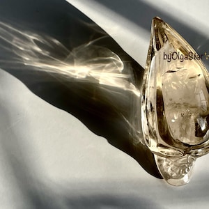 Pode incluir: Um cinzeiro de vidro âmbar em forma de gota. O cinzeiro tem uma superfície texturizada e está sobre um fundo claro. A sombra do cinzeiro é projetada na superfície. O texto "byOlgaStar" é visível.