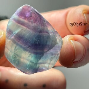 May include: A polished fluorite gemstone with a mix of purple, blue, and green hues. The stone has a smooth, rounded shape and is held in a person's hand. The text "byOlgaStar" is visible in the image.