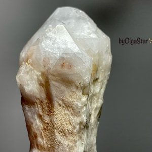 Pode incluir: Uma aproximação de um cristal de quartzo transparente com uma ponta. O cristal tem uma superfície áspera e texturizada com áreas bege e laranja claro. O fundo é cinza desfocado. O texto "byOlgaStar" é visível.