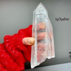 Può includere: Un cristallo di quarzo trasparente con una punta e una superficie ruvida e strutturata. Il cristallo è tenuto contro uno sfondo grigio, con una manica rossa a maglia visibile. Il cristallo è una gemma naturale e grezza.