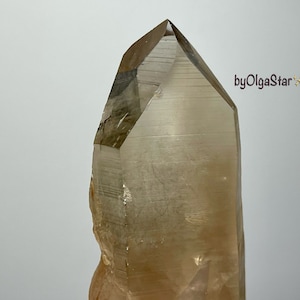 Puede incluir: Una gran punta de cristal marrón claro translúcido con una parte superior facetada. El cristal tiene una textura rugosa y natural en un lado y está sobre un fondo blanco. El texto "byOlgaStar" es visible en la esquina superior derecha.