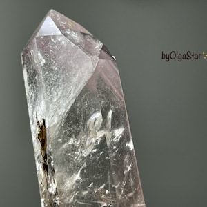 May include: A clear quartz crystal with a pointed tip. The crystal has a translucent appearance with internal fractures and inclusions. The background is a solid gray. The text "byOlgaStar" is visible in the image.