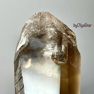 Puede incluir: Un cristal de cuarzo ahumado marrón translúcido con una superficie rugosa y texturizada. El cristal tiene un aspecto natural, sin pulir, con un interior de color claro y un exterior más oscuro, de tono ámbar. El texto "byOlgaStar" está incluido.