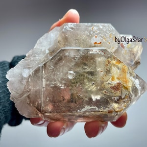 May include: A large, clear quartz crystal cluster with a hexagonal shape. The crystal has a light brown hue with areas of orange and brown inclusions. The text "byOlgaStar" is visible on the crystal.