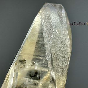 Puede incluir: Primer plano de un cristal de cuarzo transparente con una punta. El cristal tiene una superficie texturizada y un tono amarillo pálido, con inclusiones oscuras. El fondo es gris liso.