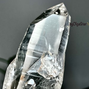 Può includere: Primo piano di un cristallo di quarzo trasparente con una punta affilata. Il cristallo è trasparente con strutture interne e imperfezioni visibili. Lo sfondo è sfocato, con un accenno di fogliame verde e il testo "byOlgaStar".